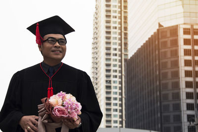 Close-up of man in graduation gown holding bouquet while standing at city