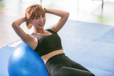 High angle portrait of smiling young woman with hands behind head exercising on medicine ball in gym