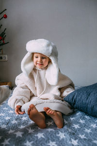 Cute child y on the bed in cozy adult clothes and a hat with earflaps.