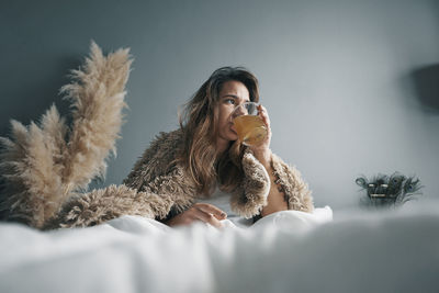 Young woman drinking herbal tea while sitting on bed against wall
