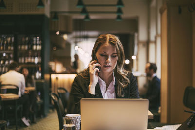 Businesswoman talking on phone while looking at laptop in restaurant