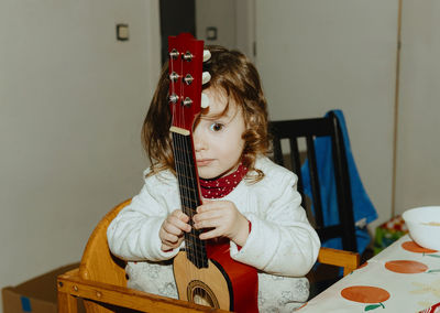 A little girl holds a guitar while sitting at the table.