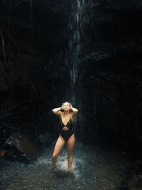 Young woman in bikini standing under waterfall in forest