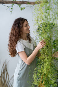 Happy young woman in dress touching houseplant at home garden, taking care. greenhouse, urban jungle