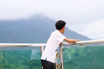 Side view of mid adult man looking at mountain while standing by railing against cloudy sky