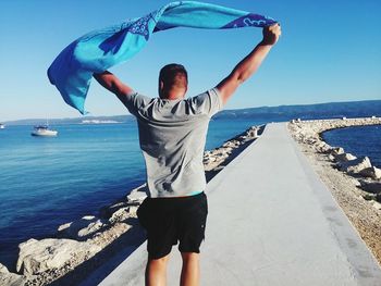 Rear view of man with towel standing on pier at beach against clear blue sky