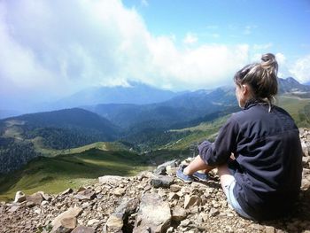 Rear view of woman relaxing on mountain against cloudy sky over landscape