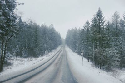 Snow covered road amidst trees against sky in winter