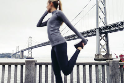 Side view of female athlete stretching leg on footpath with oakland bay bridge in background