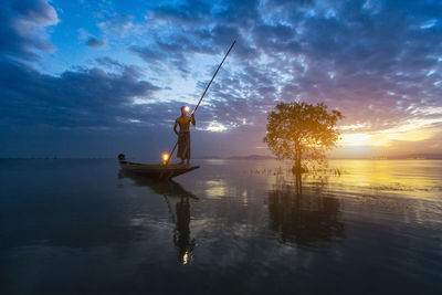 Silhouette man standing in boat on sea against cloudy sky during sunset