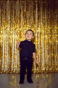 Boy in black clothes at christmas standing in a room on a background of gold tinsel on the wall