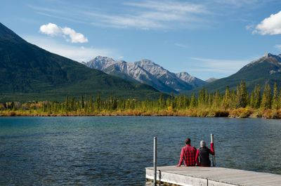 People sitting at lake by mountains against sky