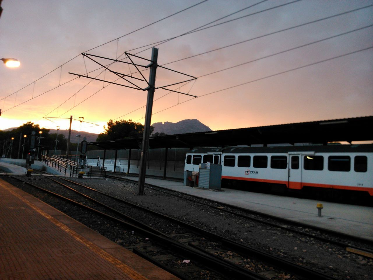 railroad track, transportation, rail transportation, railroad station platform, public transportation, power line, mode of transport, railroad station, electricity pylon, sky, train - vehicle, sunset, travel, train, cable, power supply, passenger train, electricity, dusk, road