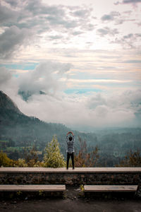 Rear view of young man standing on retaining wall against cloudy sky