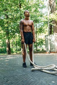 Full length of shirtless man holding rope while standing against trees