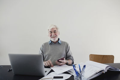 Portrait of smiling senior man with laptop and bills sitting against white wall