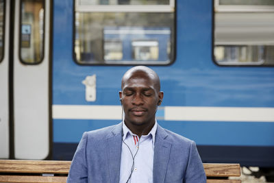 Bald commuter listening music while sitting with eyes closed against subway train at railroad station