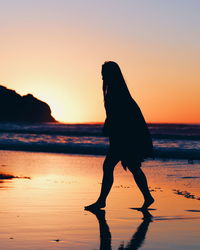 Silhouette woman walking at beach against sky during sunset