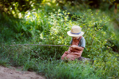 Cute european boy in hat and overalls in summer with stick fishing rod fishes in the grass near 