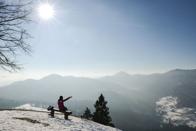 Woman pointing while sitting on bench at snowcapped mountain