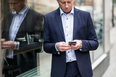 Contemplating mature businessman using smart phone while standing by glass window