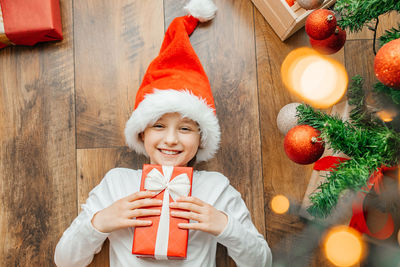 Happy teenage girl in santa hat lies on floor next to  christmas tree with a gift box in her hands