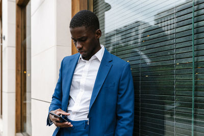 Young male entrepreneur using smart phone while leaning on window in city