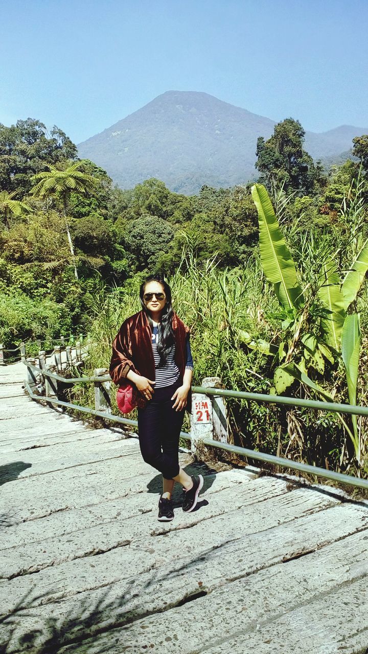 full length, casual clothing, lifestyles, leisure activity, tree, person, mountain, clear sky, looking at camera, standing, portrait, young adult, nature, smiling, day, sunlight, plant, front view