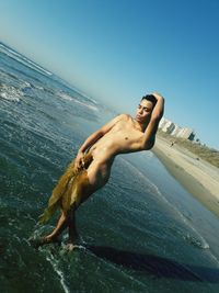 Naked young man posing at beach against clear sky