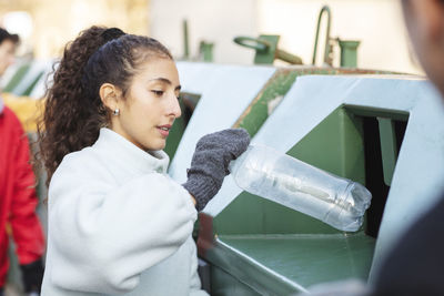 Smiling female environmentalist with friend throwing plastic bottle in garbage can