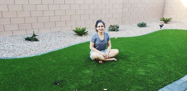 Full length portrait of smiling woman sitting on turf against wall