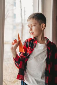 A boy with an orange paper airplane, sits at the window and looks out into the street.