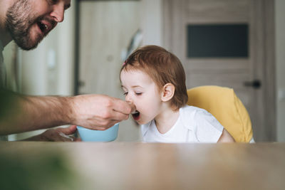 Happy father young man feeds baby girl little daughter in kitchen at home