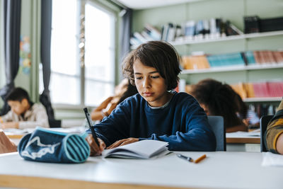 Focused boy writing in book while studying in classroom at elementary school