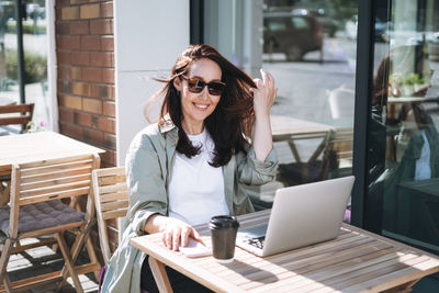 Adult smiling brunette business woman forty years in stylish shirt working on laptop in cafe at city 