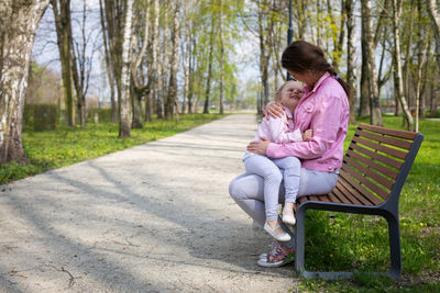In a park on a bench, a mother holds her smiling daughter on her lap.