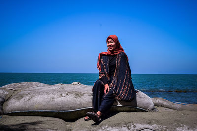 Young woman sitting by sea against blue sky