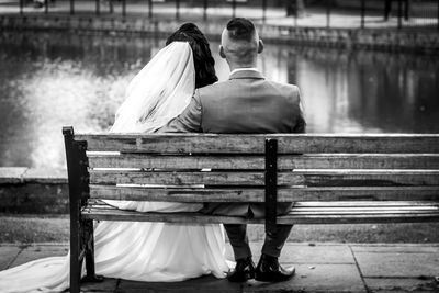 Rear view of wedding couple sitting on bench by lake