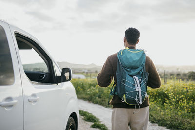 Rear view of man with backpack looking at field standing by van against sky