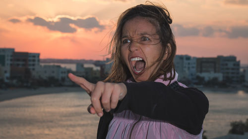 Angry woman shouting by sea in city against sky during sunset