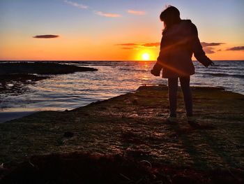 Rear view of woman with arms outstretched standing at sea shore against sky during sunset