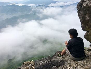 Rear view of man sitting on rocky mountain during foggy weather