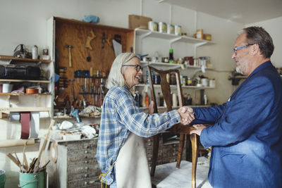 Senior owner shaking hands with customer while selling wooden chair at workshop