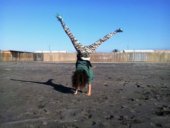 Girl doing handstand on sand at beach against clear blue sky