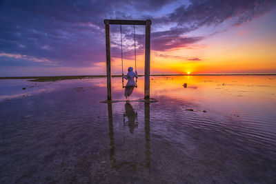 Rear view of mid adult woman swinging at beach against sky during sunset