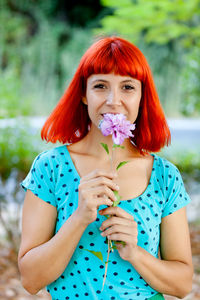 Portrait of smiling woman holding flower standing against tree