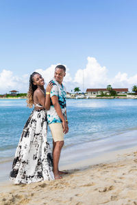 Portrait of a young smiling couple standing on the beach against the sea and clear sky