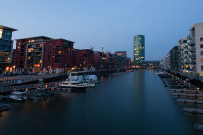 Sailboats moored on river by illuminated buildings against sky at dusk