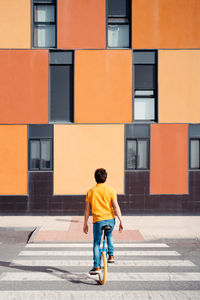 Back view of unrecognizable talented man in casual wear sitting on unicycle crossing road on zebra on modern urban street with colorful building