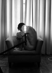 Naked man sitting on chair by illuminated light bulb at home